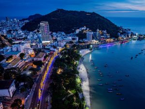 Aerial view of a coastal city at dusk with a sandy beach, waterfront lights, boats, and lush green hills by the sea.