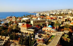 Coastal city with historic stone buildings, a blue-domed church, palm trees, and stunning Mediterranean Sea views.