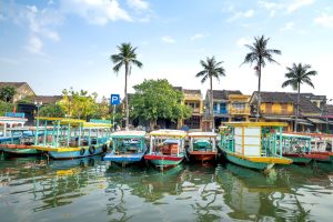 Colorful wooden boats docked along a peaceful riverside with palm trees and traditional buildings under a clear blue sky.