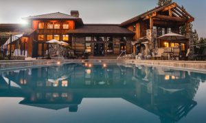 Luxury rustic lodge with a swimming pool, outdoor seating, stone accents, and warm lighting at sunset.