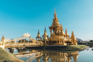 Golden temple with ornate architecture, decorative bridge, and reflective pond under a clear blue sky.